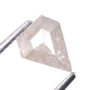 May include: A light-colored, kite-shaped diamond held by metal tweezers. The diamond has a geometric cut with a central, open facet. The stone's surface has a slightly textured appearance, and it is set against a white background.