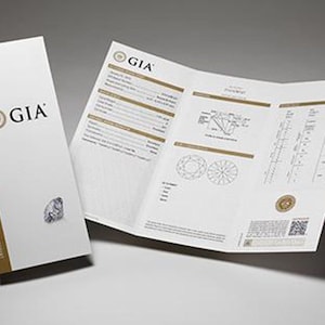May include: A GIA Diamond Grading Report and accompanying booklet. The booklet is white with gold accents and the GIA logo. The report is open, displaying diamond details and diagrams. The background is a light gray.