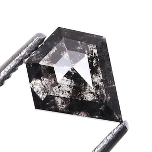 Peut inclure: Un diamant taillé en bouclier avec un motif moucheté gris foncé et noir. La pierre précieuse est maintenue par une pince argentée sur fond blanc. La forme et la couleur uniques du diamant sont mises en valeur.
