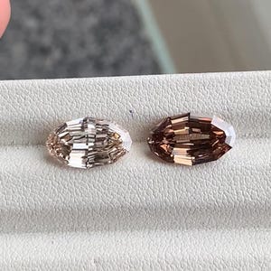 Pode incluir: Duas pedras preciosas ovais facetadas. Uma é de cor champanhe clara, e a outra de um tom castanho avermelhado mais escuro. As gemas são exibidas em uma superfície branca, mostrando sua lapidação. Ideal para a criação de joias.