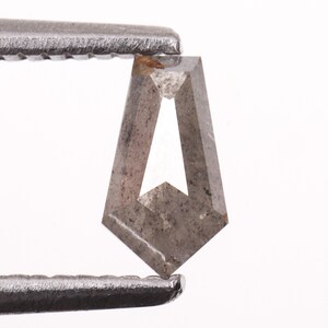Könnte beinhalten: Ein grauer, schildförmig geschliffener Diamant, der von einer Metallpinzette gehalten wird. Der Diamant hat eine einzigartige Form mit spitzem Boden und trapezförmiger Oberseite, mit einem weißen, dreieckigen Inneren. Der Stein wird vor weißem Hintergrund gehalten.