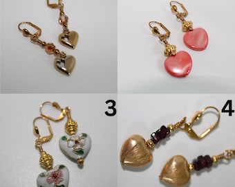 May include: Four pairs of heart-shaped earrings. The first pair has gold-colored hearts and amber-colored beads. The second pair has pink, pearlescent hearts. The third pair has white hearts with floral designs. The fourth pair has gold hearts and red beads.