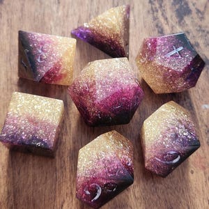 May include: Set of seven polyhedral dice with a glittery, two-tone design. The dice feature a gradient of gold, purple, and black. Each die has white numbering. The dice are arranged on a wooden surface.