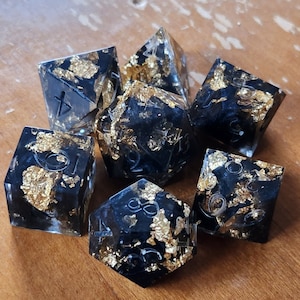 May include: Set of seven polyhedral dice with a black base and gold leaf inclusions. The dice are transparent with white numbering. The dice are arranged on a wooden surface.