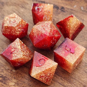 Può includere: Set di sette dadi poliedrici nei toni del rosso e dell'oro. I dadi sono realizzati in un materiale traslucido con inclusioni di glitter, ognuno con un numero diverso di facce. I numeri sono incisi in un colore a contrasto.