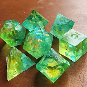 May include: Set of seven polyhedral dice in various shapes, including a cube, a d4, and a d20. The dice are transparent green with iridescent inclusions, and the numbers are etched in a lighter green. The dice are designed for tabletop role-playing games.