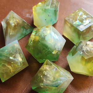 Könnte beinhalten: Sieben polyedrische Würfel aus Harz. Jeder Würfel hat eine andere Form und weist grüne und gelbe Farbtöne auf. Einige Würfel enthalten Goldflocken und sichtbare Zahlen.