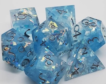 May include: A set of seven blue resin dice with iridescent glitter inclusions. Each die has black numbering. The dice are in various geometric shapes, including a d4, d6, d8, d10, d12, d20, and a percentile die.
