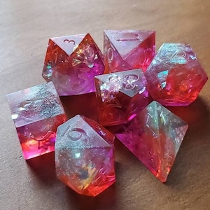 May include: Set of seven polyhedral dice in shades of red, pink, and orange. The dice are transparent with iridescent inclusions, and the numbers are visible. The dice are arranged on a wooden surface.