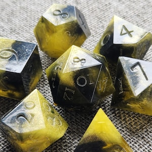 May include: A set of seven polyhedral dice for tabletop games. The dice are a mix of black and yellow resin with silver numbering. The dice include a d4, d6, d8, d10, d12, d20, and a percentile die.