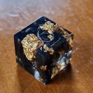 May include: A black resin die with gold leaf inclusions. The die has etched symbols and numbers on its faces. The die is sitting on a wooden surface.