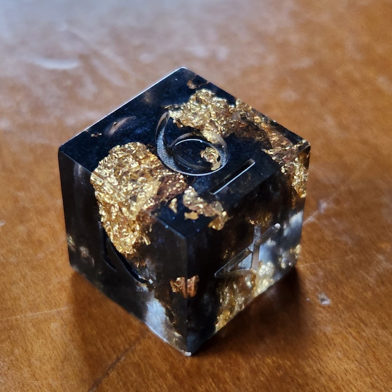 May include: A black resin die with gold leaf inclusions. The die has etched symbols and numbers on its faces. The die is sitting on a wooden surface.