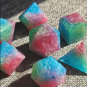 May include: A set of seven polyhedral dice in various shapes. Each die is a gradient of blue, green, and red with glitter embedded throughout. The dice are numbered with gold-colored numerals.