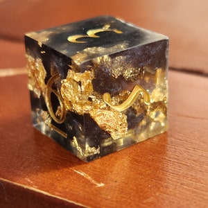 May include: A handcrafted, square resin die with gold leaf inclusions and gold-colored numerals. The die features a black interior and is set against a warm, wooden surface. The numbers are in a stylized font.