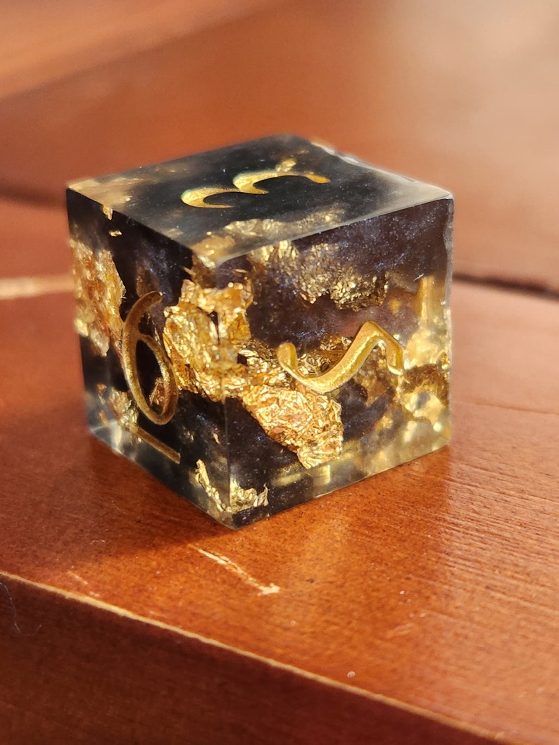 May include: A handcrafted, square resin die with gold leaf inclusions and gold-colored numerals. The die features a black interior and is set against a warm, wooden surface. The numbers are in a stylized font.