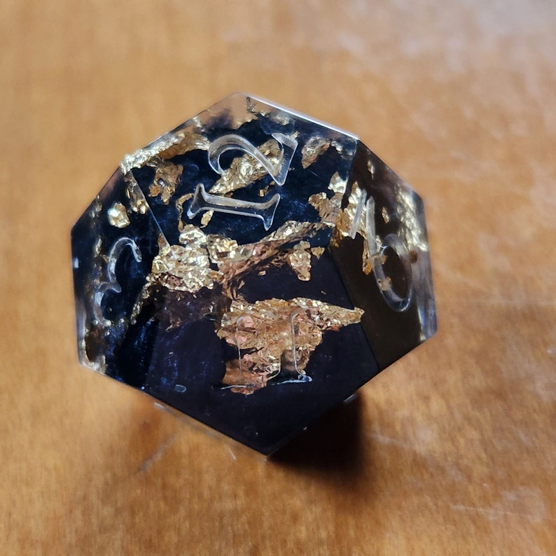 May include: A black and gold resin D20 die with clear, etched numbers. The die is filled with gold leaf flakes. The numbers are etched in a clear font. The die is a polyhedral shape, used for tabletop games.