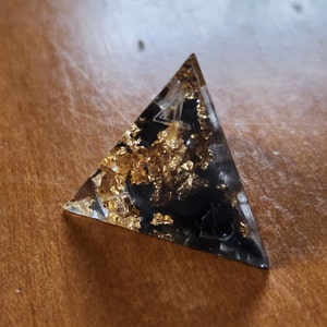 May include: A clear resin pyramid with a black base and gold leaf accents. The pyramid shape is a geometric design, and the gold leaf adds a touch of luxury. The pyramid is a decorative item.