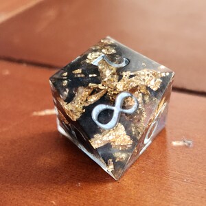 May include: A clear, square-shaped resin die with a black interior and gold leaf accents. The die features silver-colored numerals and an infinity symbol. The object is set against a brown wooden surface.