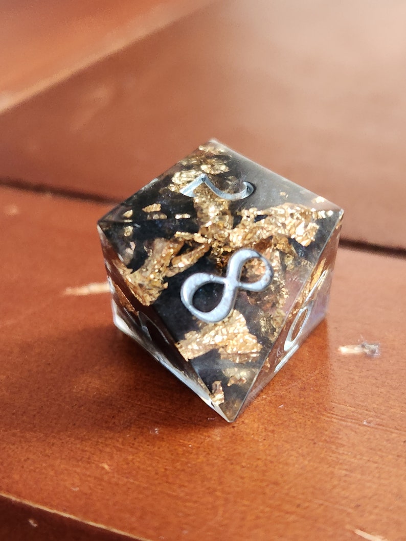 May include: A clear, square-shaped resin die with a black interior and gold leaf accents. The die features silver-colored numerals and an infinity symbol. The object is set against a brown wooden surface.