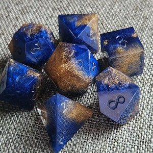 May include: Set of seven polyhedral dice in various shapes. The dice are a deep blue with gold glitter accents. Each die has white numbers or symbols. Ideal for tabletop games.