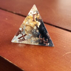 May include: A clear, triangular resin paperweight with embedded gold leaf and copper-colored Roman numerals. The paperweight is set on a wooden surface, showcasing its geometric shape and decorative elements.