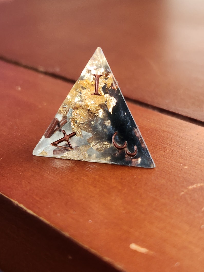 May include: A clear, triangular resin paperweight with embedded gold leaf and copper-colored Roman numerals. The paperweight is set on a wooden surface, showcasing its geometric shape and decorative elements.