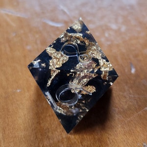 May include: A black, diamond-shaped resin object with gold leaf inclusions. The clear, raised numbers "16" are embedded on the surface. The object is likely a decorative piece or a unique dice.
