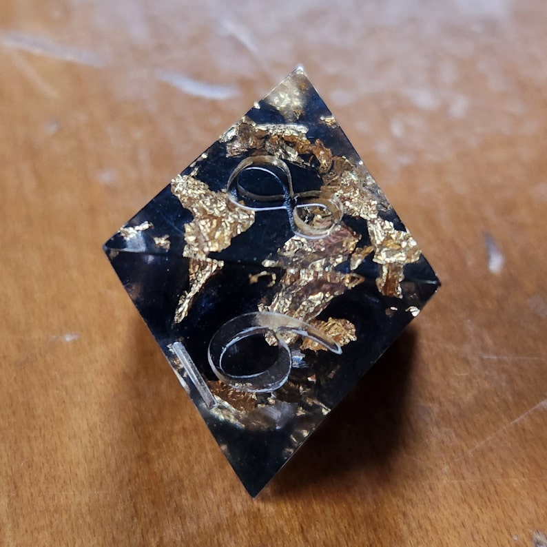 May include: A black, diamond-shaped resin object with gold leaf inclusions. The clear, raised numbers "16" are embedded on the surface. The object is likely a decorative piece or a unique dice.
