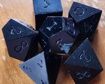 May include: A set of seven black polyhedral dice for tabletop games. Each die has engraved white numbers and symbols. The dice are arranged on a wooden surface, showcasing their various shapes and sizes.