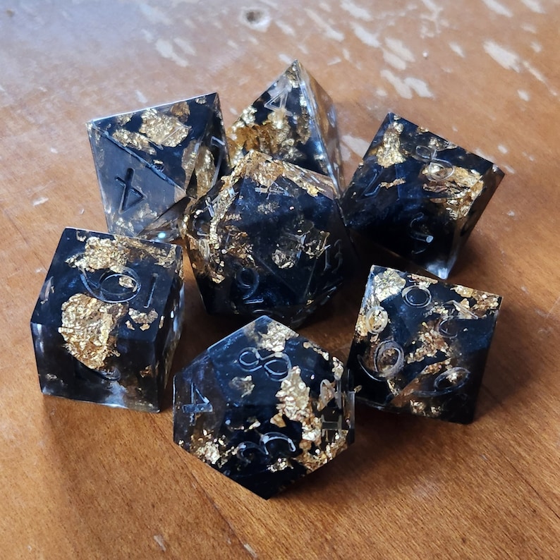 May include: Set of seven polyhedral dice with a black base and gold leaf inclusions. The dice are transparent with white numbering. The dice are arranged on a wooden surface.
