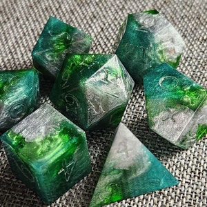 May include: Set of seven polyhedral dice in shades of green and silver. The dice are made of a translucent material, with visible swirls of color and etched numbers. The dice are designed for tabletop role-playing games.
