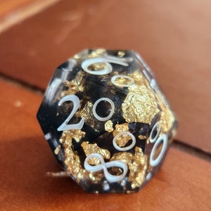 May include: A close-up of a twenty-sided die. The die is black with gold leaf inclusions and white numbering. The die is sitting on a brown surface.