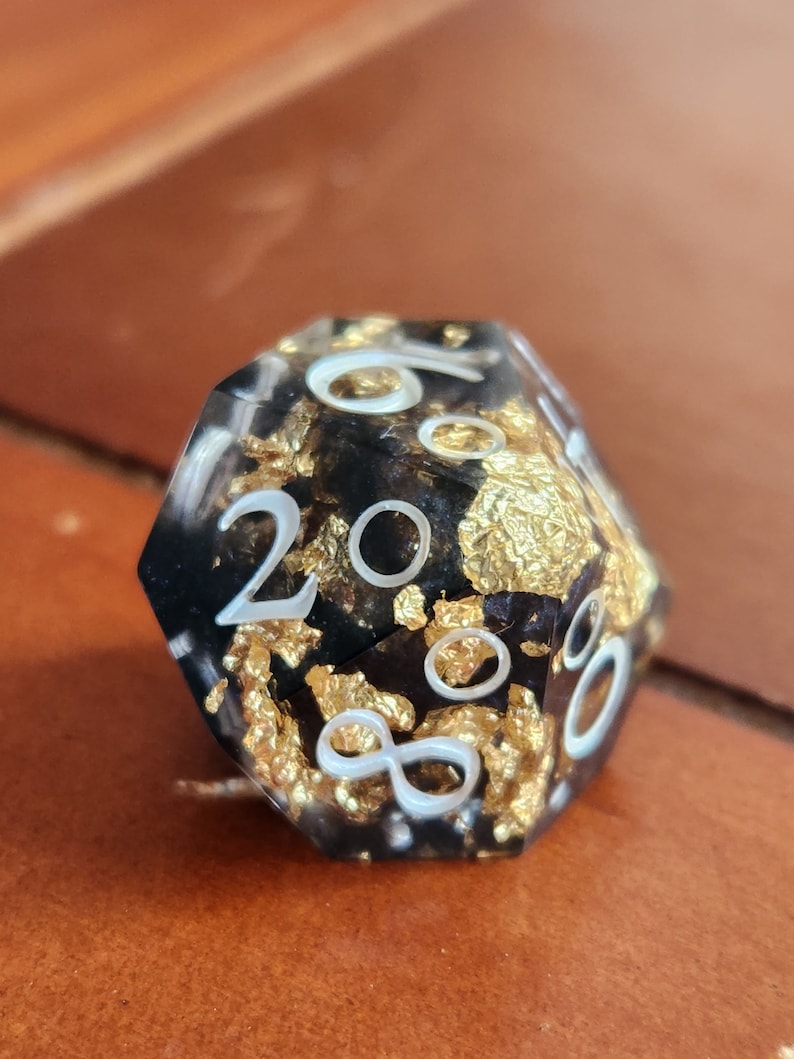 May include: A close-up of a twenty-sided die. The die is black with gold leaf inclusions and white numbering. The die is sitting on a brown surface.