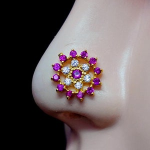 Può includere: Un anello per il naso in oro con un design circolare che presenta un fiore centrale con pietre bianche circondate da un anello di pietre rosa.