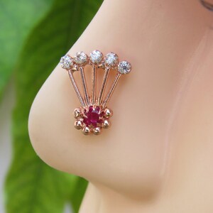 May include: A rose gold nose ring with a red gemstone surrounded by small gold beads. Five clear gemstones are attached to the top of the ring, forming a fan shape.
