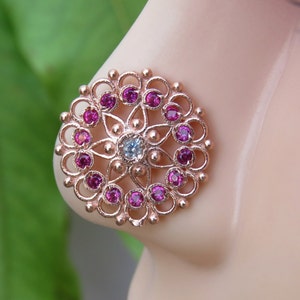 May include: A rose gold nose ring with a filigree design and pink gemstones. The center of the ring features a larger, clear gemstone.