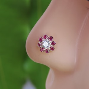 Puede incluir: Un aro de nariz de plata con una piedra blanca rodeada de piedras rojas en forma de flor.