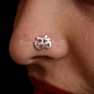 May include: A silver nose ring with an Om symbol. The ring is worn in a person's left nostril.