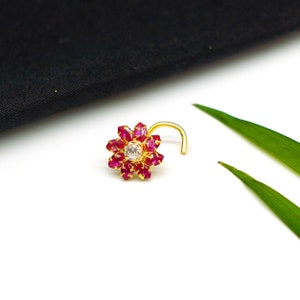 Puede incluir: Un aro de nariz de oro con un diseño de flor. La flor está hecha de pequeñas piedras rojas y una piedra blanca más grande en el centro.
