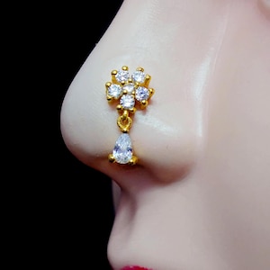 May include: Gold nose ring with a flower design and a teardrop-shaped pendant, both adorned with clear cubic zirconia stones.