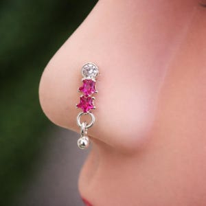 May include: A silver nose ring featuring a clear, round gemstone at the top, followed by two pink star-shaped gems. A small silver ball dangles from the bottom. The jewelry is shown on a person's nose.