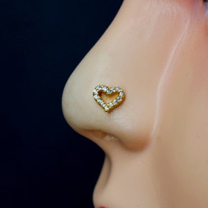 Puede incluir: Un aro de nariz de oro con un diseño en forma de corazón. El corazón está rodeado de pequeñas piedras transparentes.