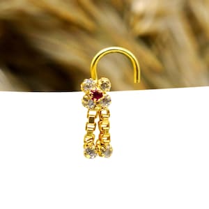 Puede incluir: Un aro de nariz de oro con una pequeña piedra roja y piedras transparentes en un diseño de flor. El aro tiene un cierre de gancho.