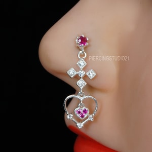 May include: A silver nose ring featuring a cross design with a heart-shaped pendant. The nose ring has a small, round, red gemstone at the top and a heart-shaped pendant with three red gemstones. The text "PIERCINGSTUDIO21" is visible.