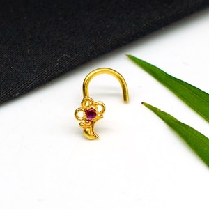 May include: A gold nose ring with a curved post and a floral design. The flower has a central red gemstone and is surrounded by intricate gold detailing. The jewelry is set against a white background with green leaves.