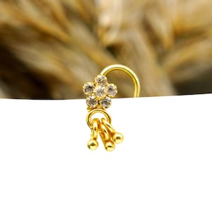 Könnte beinhalten: Ein goldener Nasenring mit einem blumenförmigen Anhänger, der mit sechs klaren, funkelnden Steinen besetzt ist. Unter der Blume hält eine kleine goldene Öse drei hängende Goldperlen, die sich frei bewegen und ein filigranes Schmuckstück bilden.