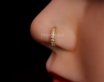 May include: A gold nose ring with a row of clear cubic zirconia stones. The ring is worn on a person's nose.