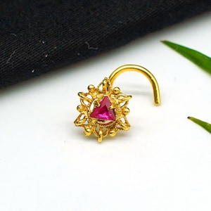 Op de afbeelding: Gouden neusring met een roze driehoekige edelsteen gezet in een decoratief filigraanontwerp.