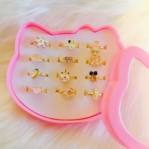 以下が含まれることがあります： 透明な蓋が付いたピンクのハート型の箱の中に、カラフルなエナメルチャームが付いた金色の指輪が12個入っています。チャームには、花、ゾウ、ロリポップ、蝶、ユニコーン、猫、ハート、花、猫、蜂、星、そして「Stay Weird」と書かれた吹き出しがあります。