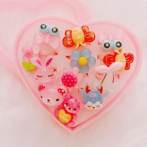 Könnte beinhalten: Eine rosa herzförmige Schachtel gefüllt mit bunten Plastikringen mit verschiedenen Tier- und Blumendesigns. Die Ringe sind für das Rollenspiel von Kindern.